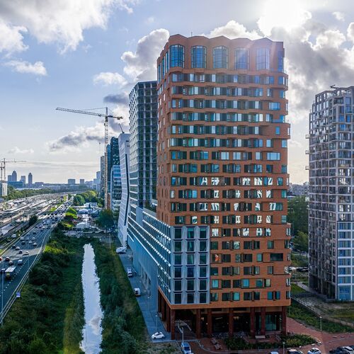 Buitenaanzicht van het project The Pulse in Amsterdam met moderne hoogbouw en stedelijke vervoersinfrastructuur.